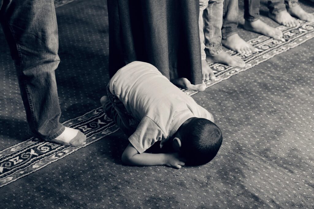 A boy kneels on the floor, reciting Islamic poetry in Urdu to an attentive group of people around him.